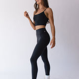 A woman models Raeya Fit's best-selling Waist Snatch leggings, designed to flatten the stomach with a double-band and 'snatch' the waist with a top rubber band. These full-length leggings feature a 25" inseam and an ultra high-waisted fit, ensuring a sleek and flattering silhouette. Made from eco-friendly fabric with post-consumer recycled content, these leggings offer both sustainability and style.