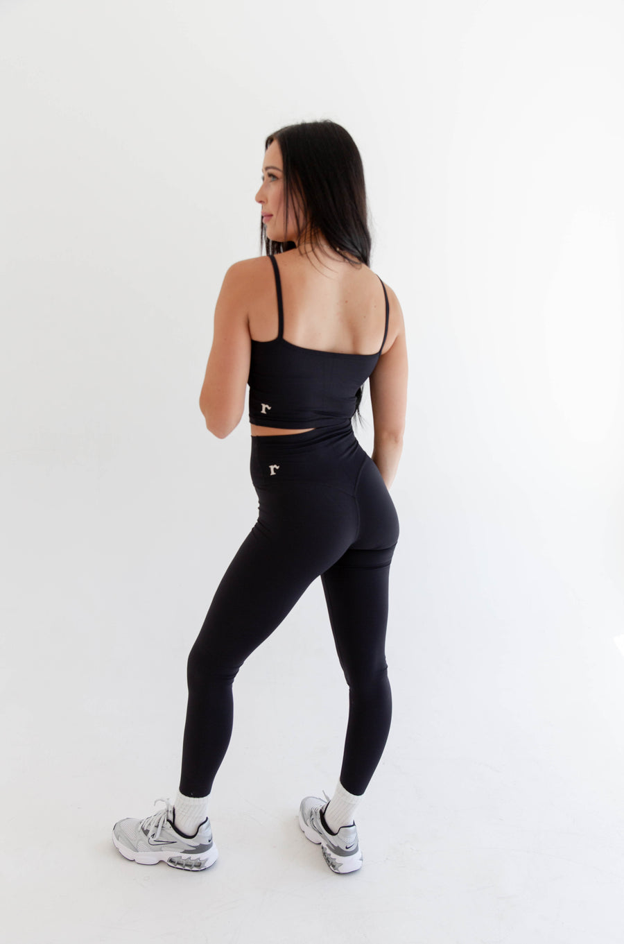 Alt text: "A woman is showcasing the best-selling Rae Butter Collection black leggings from Raeya Fit. These 5-star reviewed leggings feature a 28" inseam suitable for all heights, a Glute-Curve back seam to accentuate the booty, no front or side seams, and a high-waisted design with a double-lined hip band to ensure they stay in place. Made from soft and comfortable fabric, they offer a sleek, form-fitting look perfect for any activity."